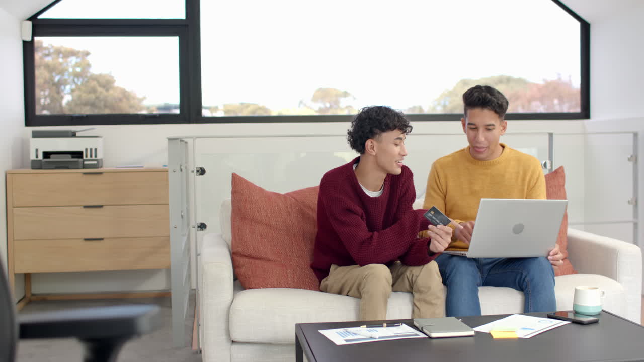 Using laptop and holding credit card, multiracial gay couple shopping online at home