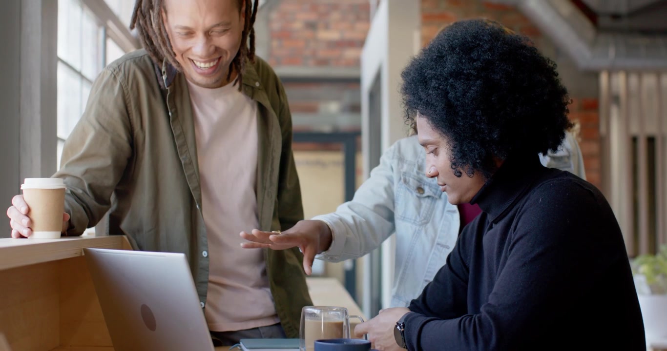 African American trio at cafe bar, peer pointing at laptop editing, seated man reaching trackpad