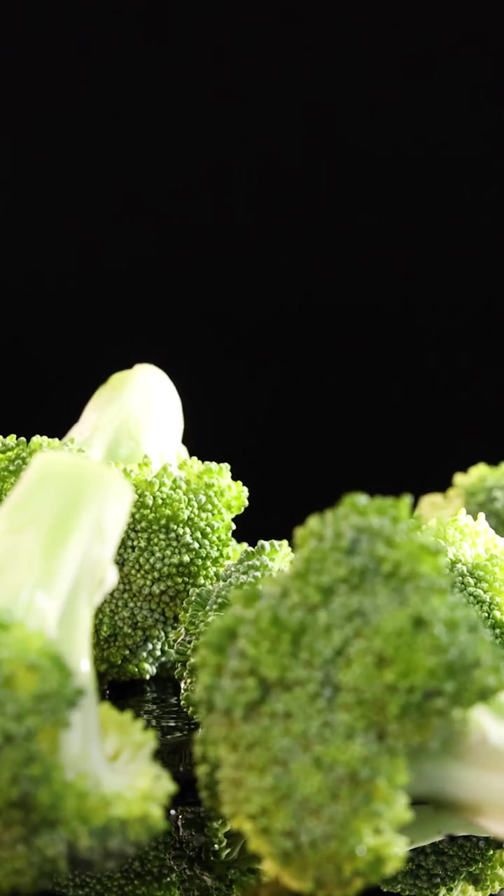 Cimette di broccoli cadono su una superficie riflettente e bagnata su uno sfondo nero, catturate in un primo piano ad alta velocità con illuminazione drammatica da studio e movimento minimo della fotocamera.