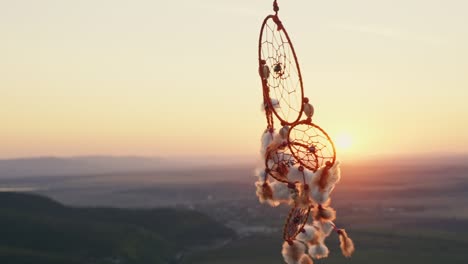 dreamcatcher with orange feathers rotate in mountains, swaying with light wind slow motion agains blue sky at sunset in summer. amulet boho style, indian style. bright disk of sun, rays of sun