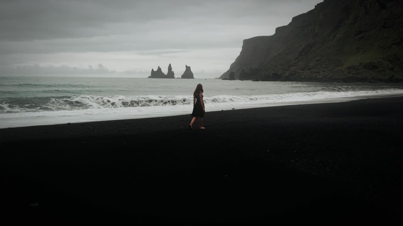 joven hermosa con vestido negro caminando por la playa de arena negra, paisaje aéreo épico del océano, islandia