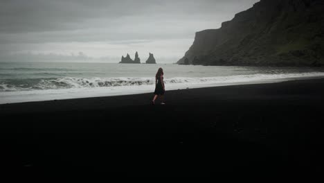 Junge-Schöne-Frau-Im-Schwarzen-Kleid,-Die-Am-Schwarzen-Sandstrand-Spaziert,-Epische-Meereslandschaft-Aus-Der-Luft,-Island