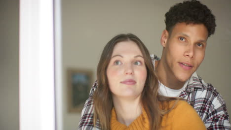 una pareja feliz y diversa mirando por la ventana y abrazándose en casa, en cámara lenta.