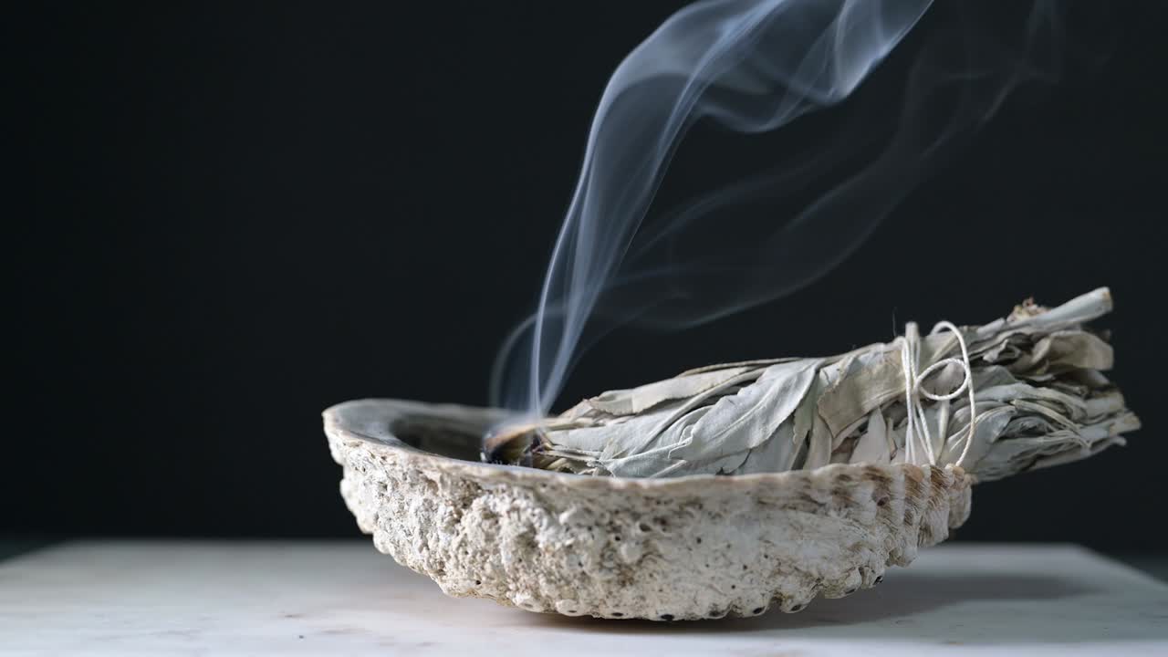 Burning sage in a shell on a wood table with black background showing sage smoke