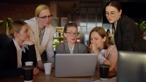 A-group-of-girls-in-business-suits-gathered-around-a-laptop-and-are-discussing-a-specific-idea-and-plan-for-working-in-a-modern-office.-Confident-girls-in-business-suits-of-different-colors-gathered-around-a-laptop-and-talked-about-a-current-topic