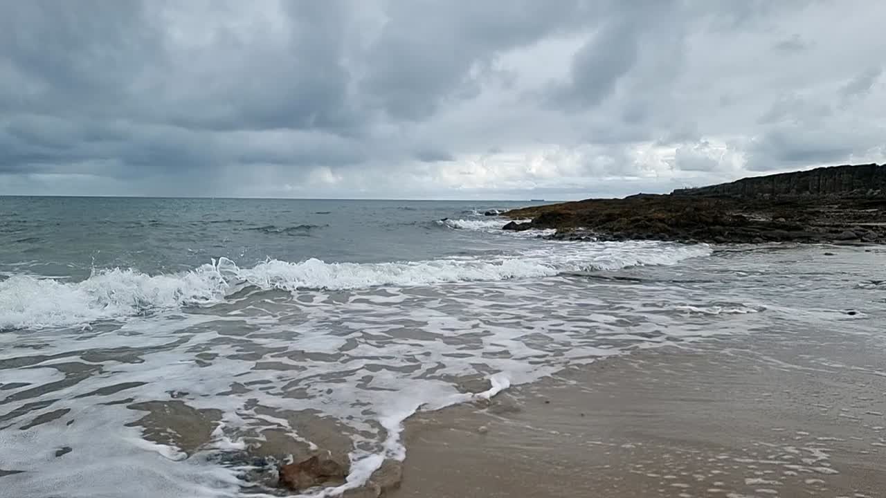 Slow motion foaming tide waves washing over sandy rugged Welsh beach coastline