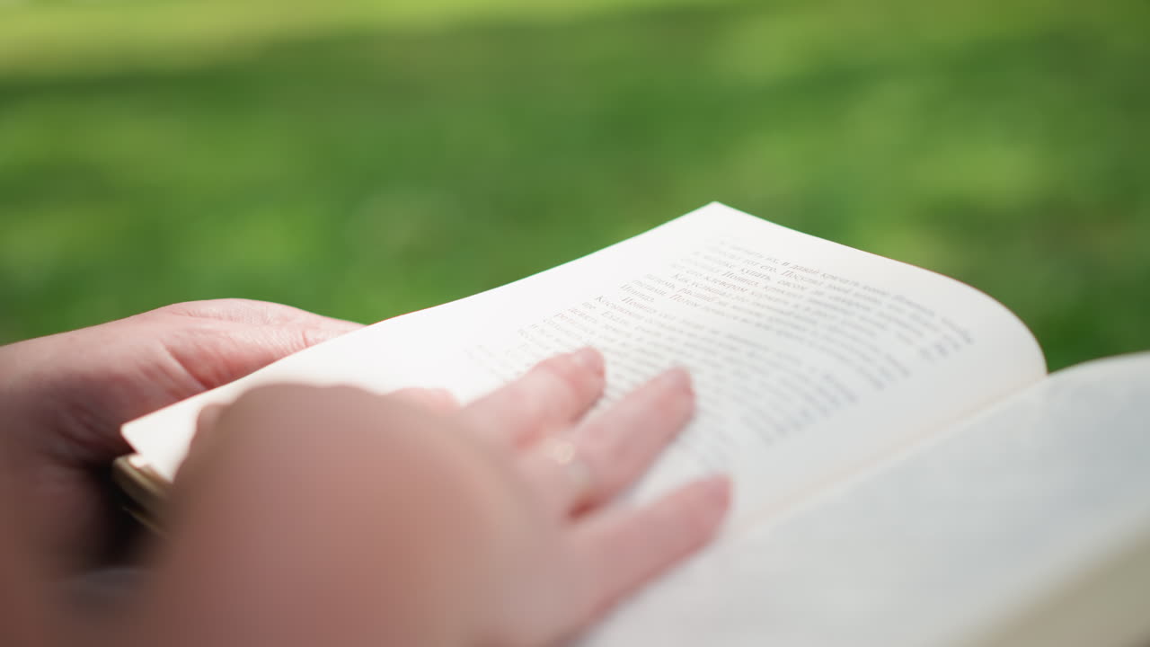 Close up of student gently flipping through open book pages while studying outdoors on green grass, soft sunlight creating calm atmosphere of concentration and learning