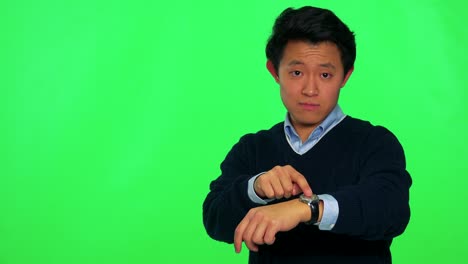 a young asian man looks at the camera and taps at his wristwatch - green screen studio