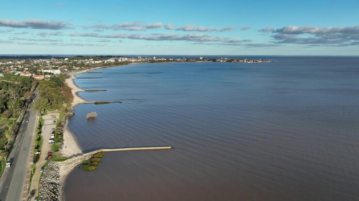 Ripresa aerea in avanti di Colonia del Sacramento lungo la costa curva della spiaggia con frangiflutti e lo skyline della baia oltre