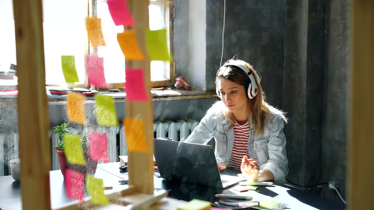 una chica rubia está escuchando música y trabajando con una computadora portátil en una oficina de loft agradable. está tomando notas en el bloc de notas, sonriendo y bailando. relajándose en el concepto de lugar de trabajo.