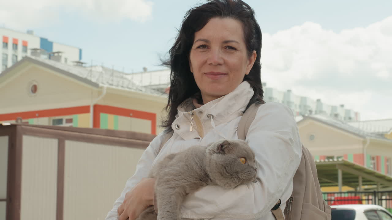 White Woman Holding Cat Near Colorful Houses, Front Porch Backdrop With Midday Light, Casual Bag Over Shoulder, Windblown Hair And Friendly Gaze, Suburban Commuter Pausing On Walk, Adoption Advocate
