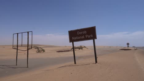 signo de nombre para el parque nacional de dorob al lado de un viejo signo roto, arena soplando en el suelo en un día soleado