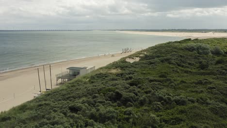 Luftaufnahme,-Die-An-Einem-Bewölkten-Tag-über-Grüne-Dünen-Und-An-Einem-Strandpavillon-Vorbei-In-Richtung-Meer-Fliegt