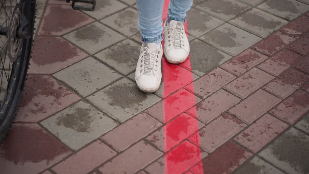 Sneakers bianche che camminano sulla linea rossa, donna in jeans chiari che cammina su un marciapiede piastrellato bagnato accanto al pedale di una bicicletta, primo piano dei piedi, atmosfera urbana piovosa, passeggiata solitaria, guida di confine, stile di vita spontaneo