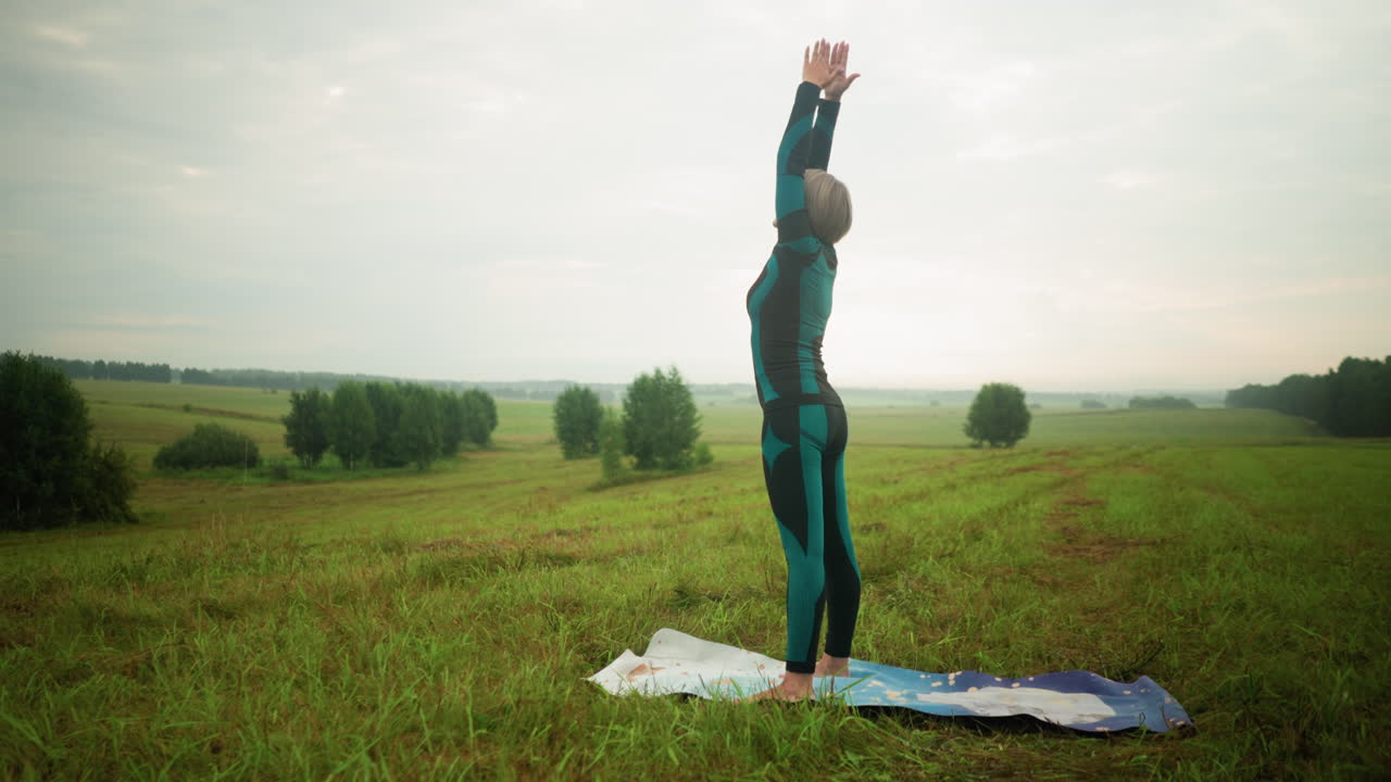 mujer de mediana edad de pie en una alfombra de yoga con los brazos extendidos levantados hacia el cielo, preparándose para una postura de flexión hacia adelante en un campo de hierba brumosa, bajo un cielo nublado con árboles alineados en la distancia