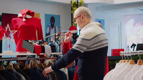 cliente senior navegando por las estanterías de la tienda de ropa decorada con xmas con el marido en la juerga de compras de navidad. pareja de edad en busca de blazer elegante como regalo para el hijo durante la temporada de vacaciones de invierno