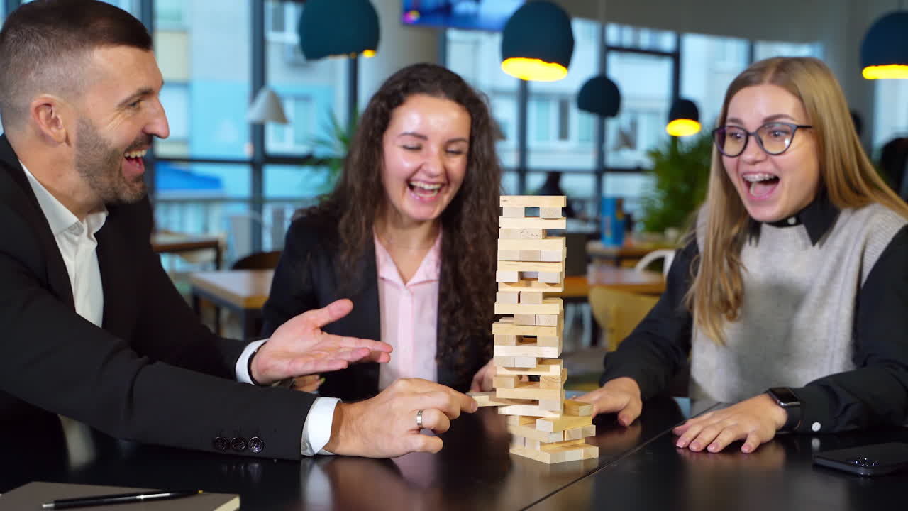 Office staff having free time at work. Teammates playing jenga sitting at table. Bearded man pulls the brick and breaks the tower.