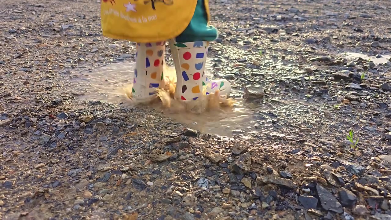 A child in colorful boots taps a puddle, then steps away in warm sunlight.