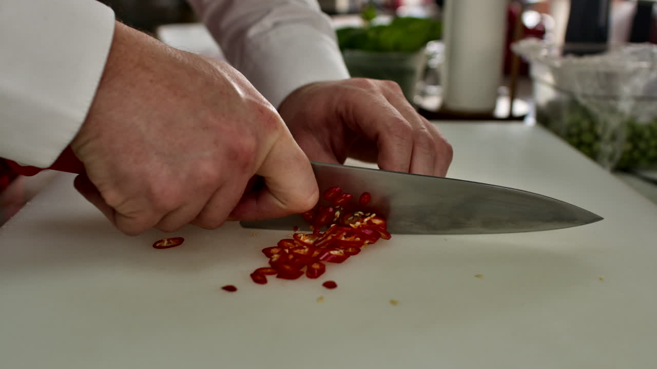 Close-up of a chef's hands finely slicing a red chili pepper. Professional culinary preparation for a spicy recipe and food styling