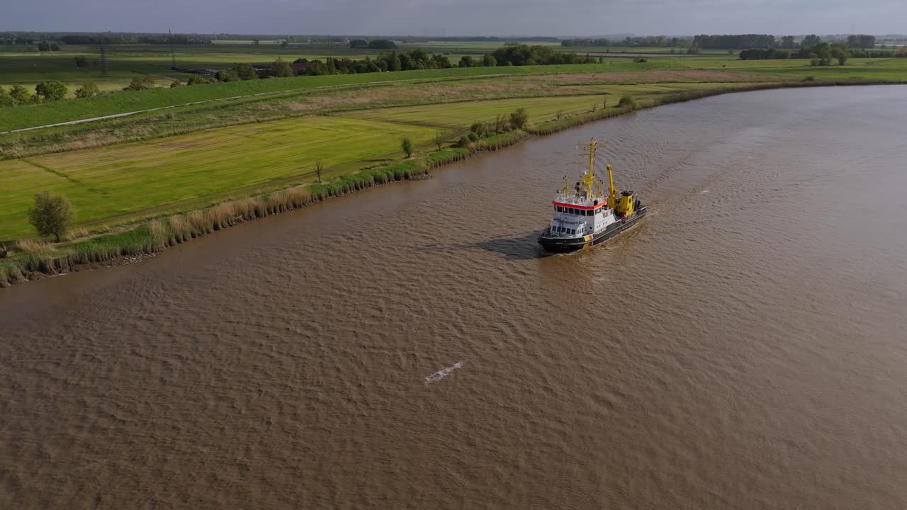 Drone footage of a utility or service vessel navigating the Ems River in Northern Germany. The camera tracks the boat while flying backward, capturing surrounding rural fields and water surface.