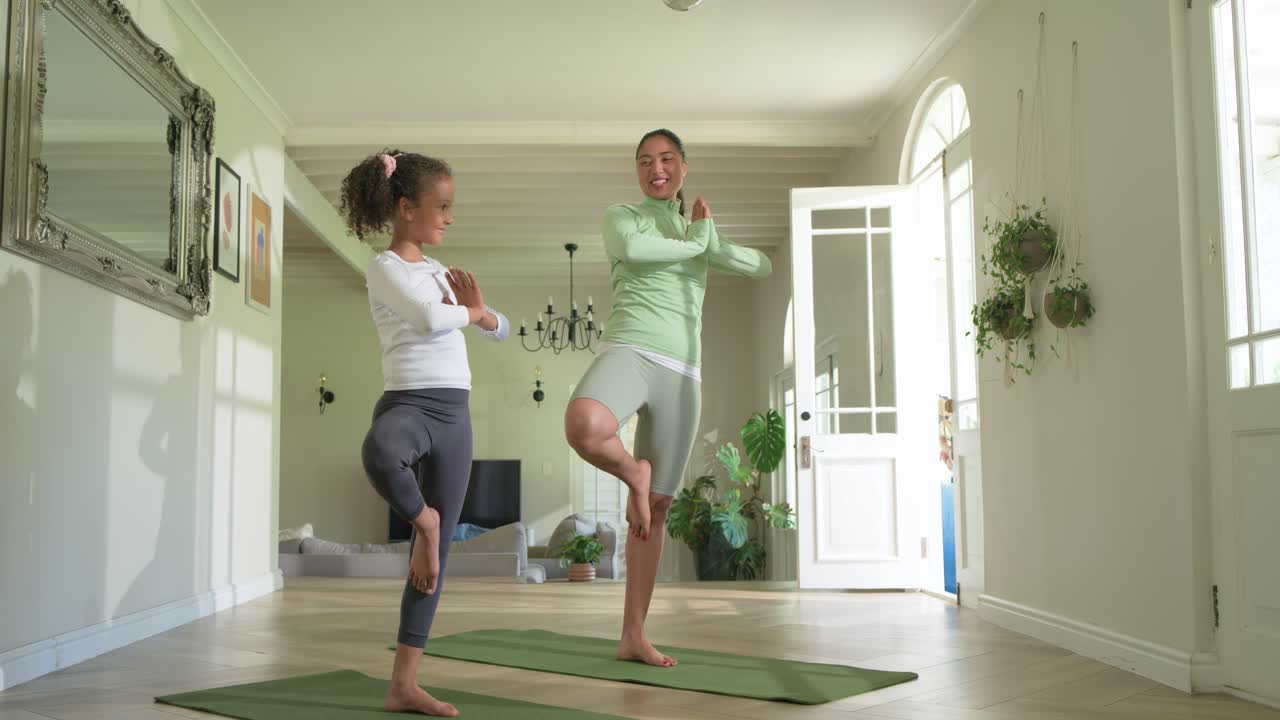 Mulher asiática guiando criança afro-americana em ioga em casa, equilibrando-se em tapetes verdes para fortalecer o vínculo.
