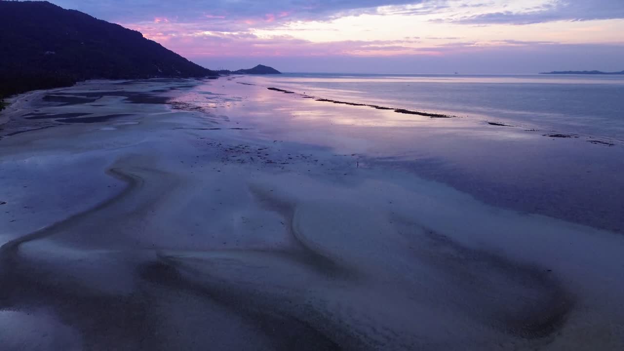 Beautiful aerial footage of sunrise over Koh Phangan in Thailand with calm ocean and pink sky colors reflecting on the beach during low tide