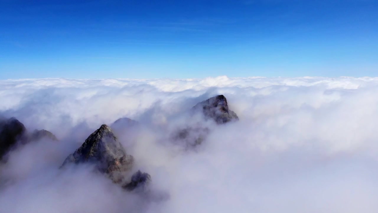 Ampio paesaggio montano che mostra cime scure e frastagliate che perforano spesse nuvole bianche sotto un cielo azzurro limpido, una vista alpina drammatica con nebbia, texture rocciosa, orizzonte aperto e natura selvaggia e silenziosa, con montagne e vette.