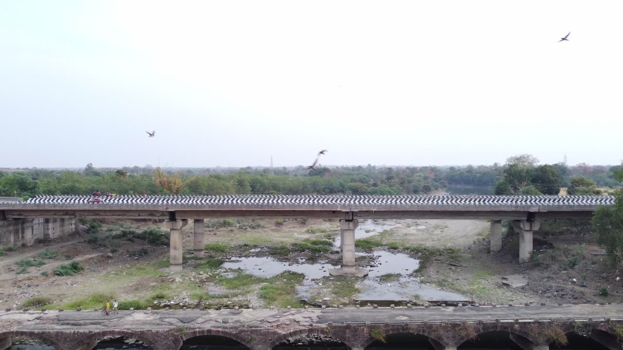 Drone charging towards a demolished old bridge in foreground with a newly built 6 lane highway bridge in background over a typical dried up river basin in the mainland Indian rural terrain
