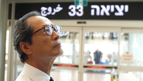 a japanese businessman stands at ben gurion airport in israel, watching the arrivals and departures board, with an exit sign in hebrew in the background