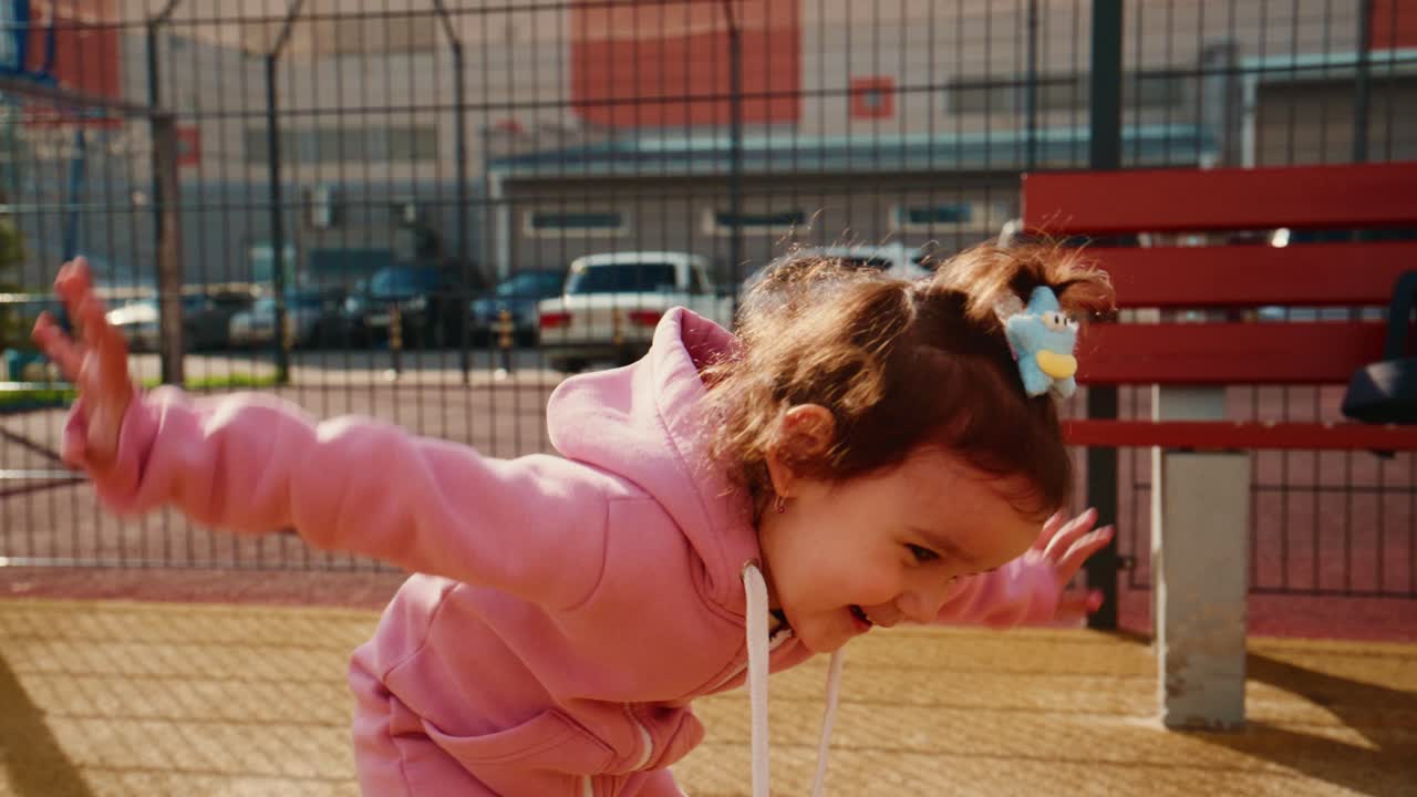 A Joyful Young Girl in a Pink Hoodie Playing on a Sports Court with Her Hair in Curls, Expressing Happiness and Energy in the Late Afternoon Sunlight