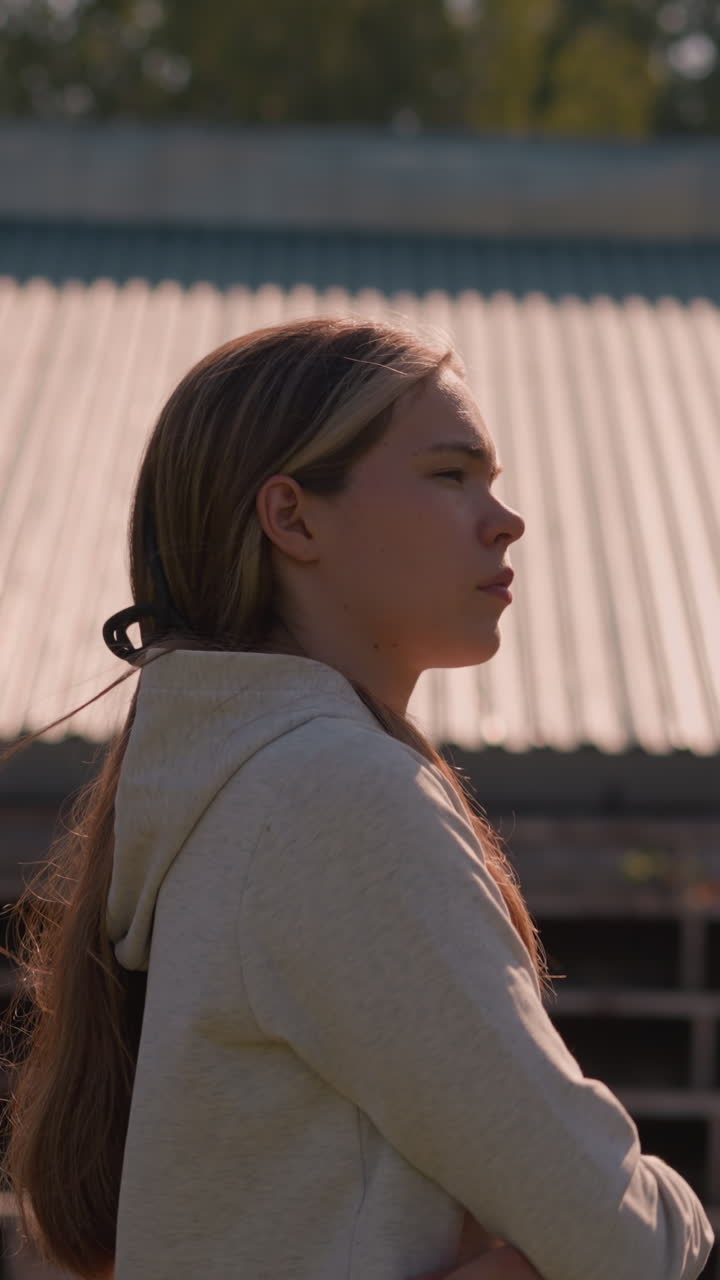 chica con capucha con el cabello largo atado hacia atrás caminando al aire libre, mirando pensativo tocó su mejilla mientras su cabello revoloteaba suavemente en la brisa, con el techo y los árboles en el fondo