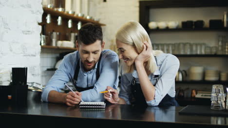 Attractive-Cafe-Workers-Standing-At-The-Bar-And-Thinking-About-New-Menu,-Man-Writing-With-Pencil