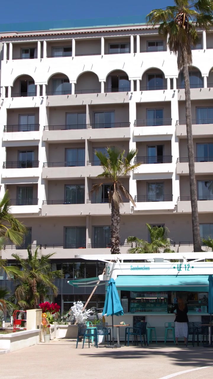 Pareja caucásica camina por un soleado paseo marítimo junto a moderna arquitectura de resort y altas palmeras.