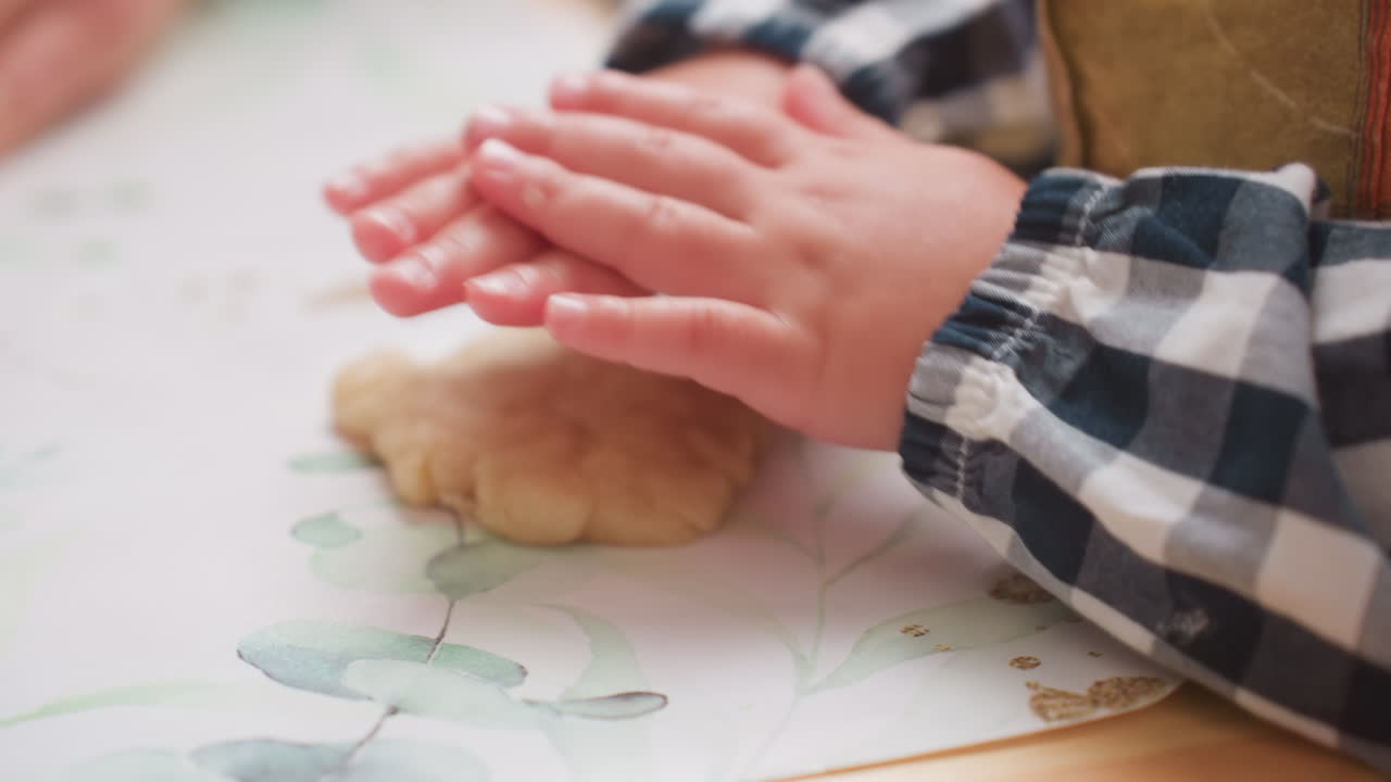 Hand view of kid in checkered sleeve pressing dough on floral table mat during playful cooking activity, small hands engaged in shaping texture with focus, showing early childhood learning