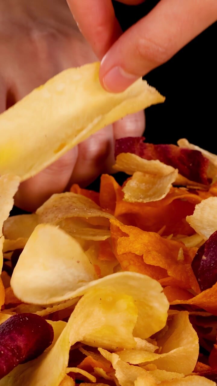 Hand selects crispy mixed root vegetable chips under studio lighting, close-up, black background, static camera