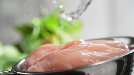 lavando pecho de pollo crudo en un colander bajo una corriente de agua salpicando en cámara lenta