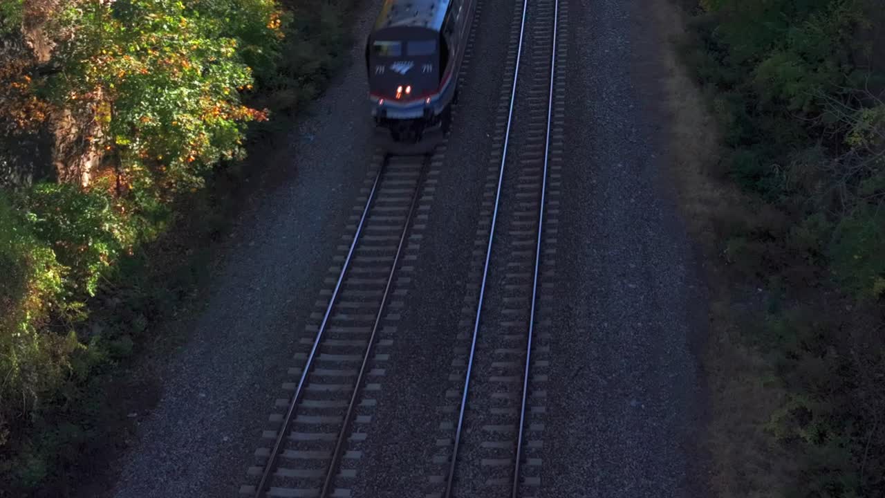 An aerial view of an Amtrak train traveling in a ravine in the Hudson Valley on a sunny day in autumn. The camera is stationary, tilted down showing colorful trees in the sunshine and in the shadows.