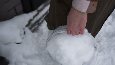gardener packing snow with bare hands into bucket while stepping through snowy threshold of rustic wooden house, door closing behind, footprints in crisp white drifts under overcast winter sky