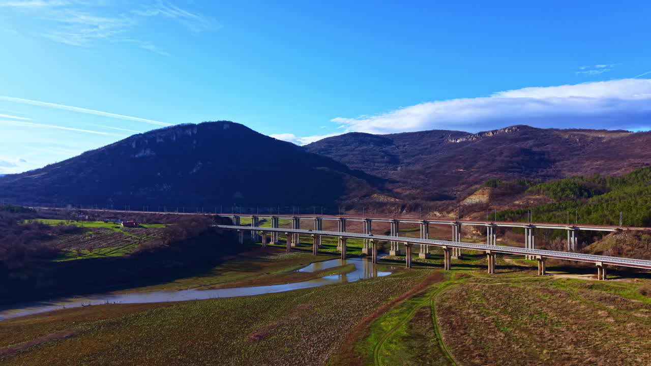 Bro krydser flod under klar himmel nær bakker og bjerge i løbet af dagen i et landligt område med åbne marker og grønne områder