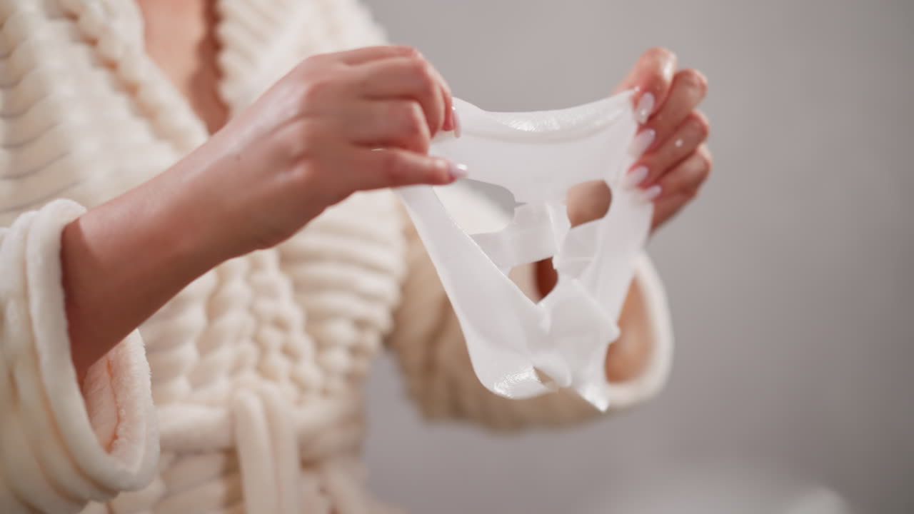 mujer sostiene una máscara facial con consistencia líquida en las manos. mujer con manicura en bata de baño a punto de ponerse una máscara de tela facial de pie en el baño