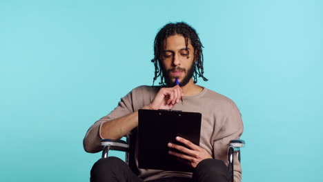 Man-in-wheelchair-writing-on-clipboard-with-pen,-studio-background