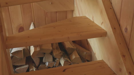 closeup leg view of person descending wooden staircase in warm cabin setting, wearing jeans and slippers, with chopped firewood stored underneath