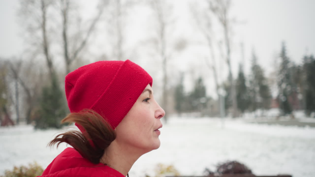 은 베니와 후디를 입고 눈 인 공원 경로를 따라 조하는 피곤한 여자의 가까운 모습, 얼어붙은 겨울 나무와 멀리 눈 먼지가 묻은 주민