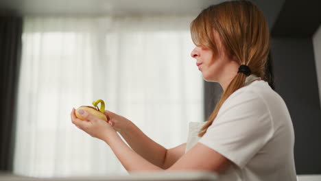Side view of young lady holding peeler and potato in both hands with hair resting on her chest as natural light softly filters through translucent curtain in modern kitchen
