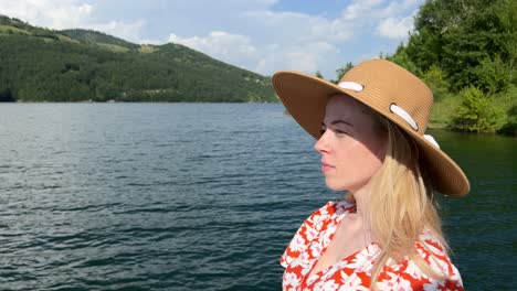 blonde girl in a straw hat stands gracefully by a lake, sunbathing and gazing at mountains and trees on a sunlit summer day