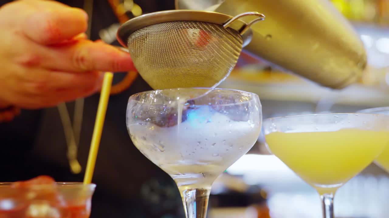 Chilled margarita being poured into a glass at a bar setting, with another cocktail nearby
