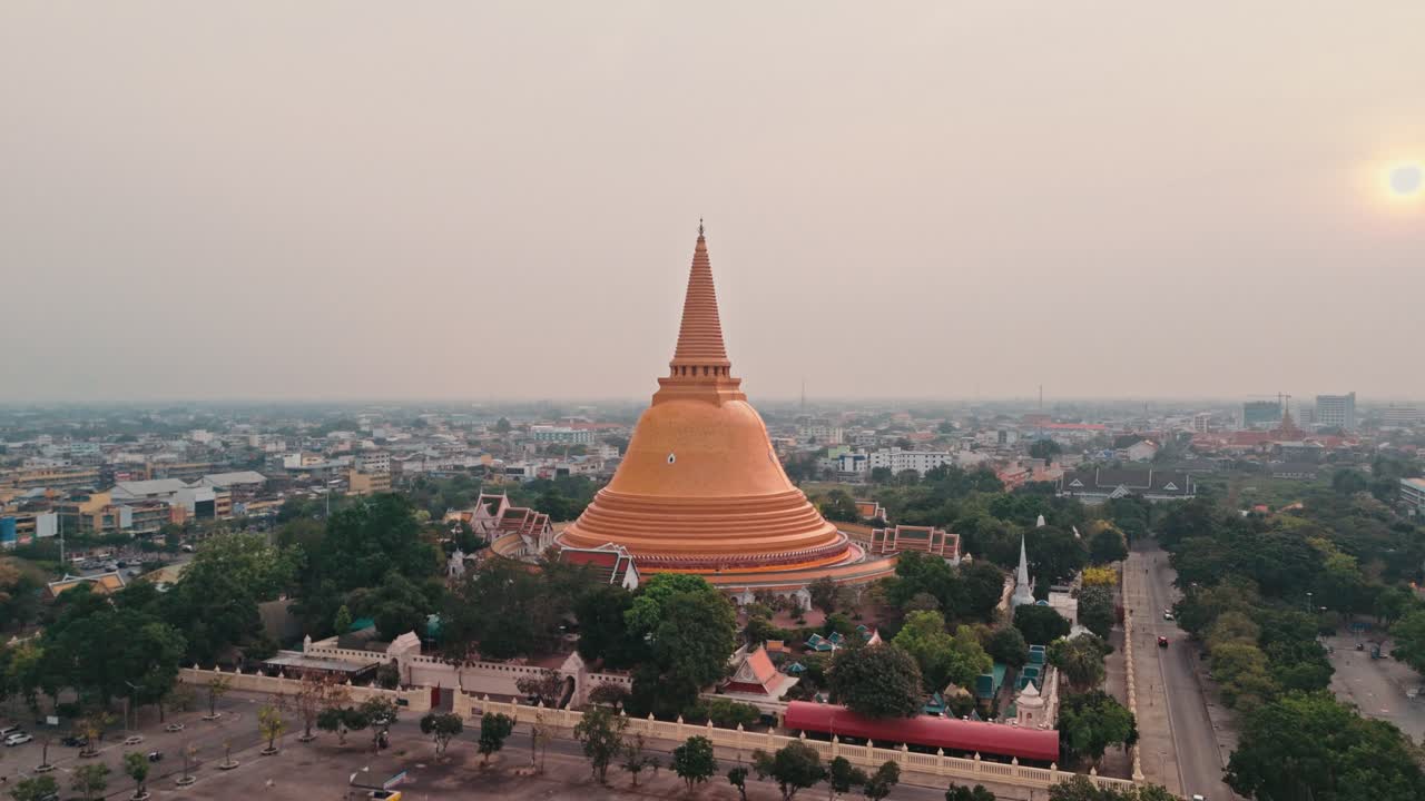 Un volo fluido di drone avanza verso un'enorme stupa dorata che si erge sopra il centro urbano, mentre un maestoso monumento buddista domina lo skyline della città in Thailandia sotto un cielo grigio e velato con il sole mattutino splendente
