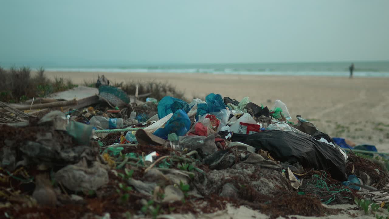 Pile of plastic waste on a beach with blurred person walking in the background, highlighting pollution