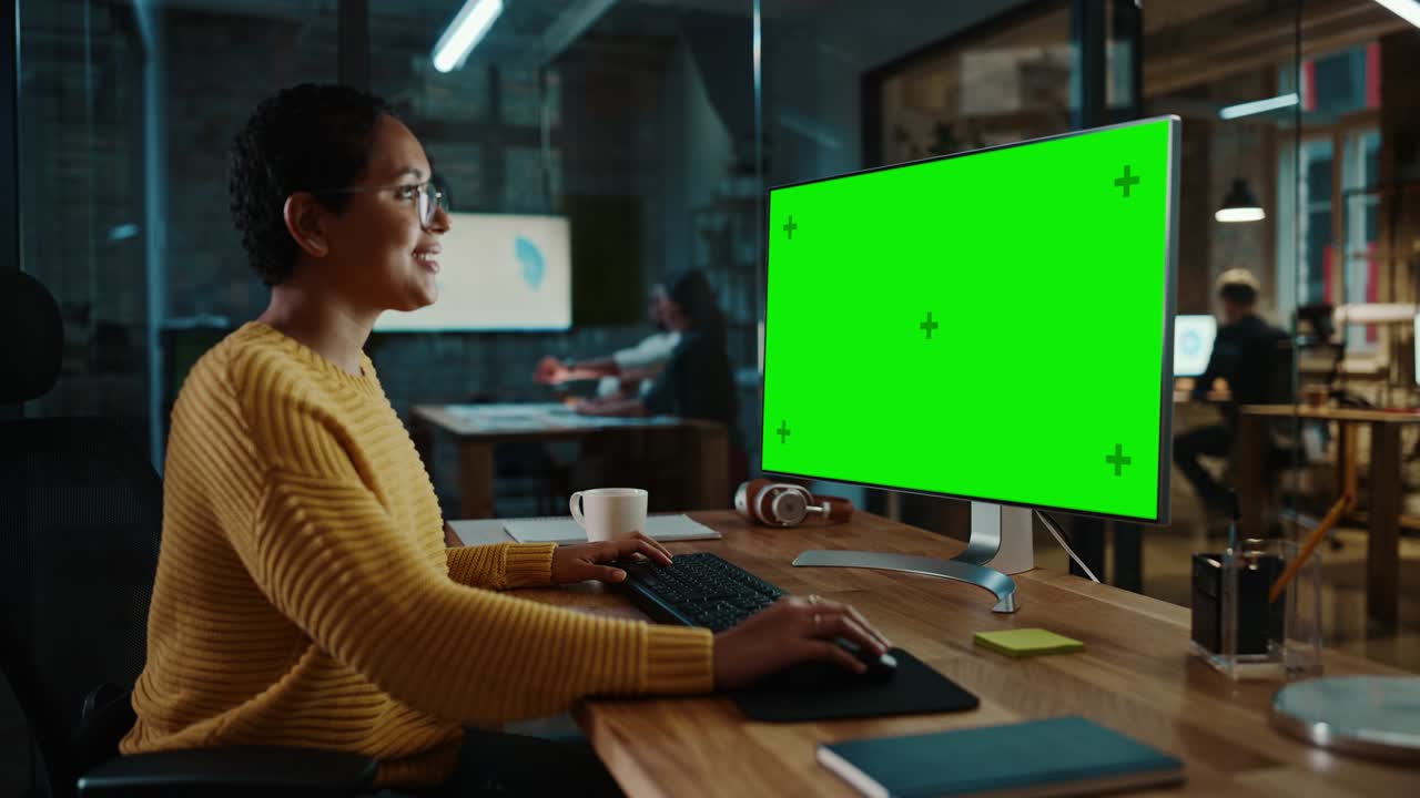 joven especialista multiétnico trabajando en una computadora de escritorio con pantalla verde simulacro de visualización en la oficina creativa. hermosa gerente femenina diversa con cabello corto y gafas está usando un jumper amarillo.