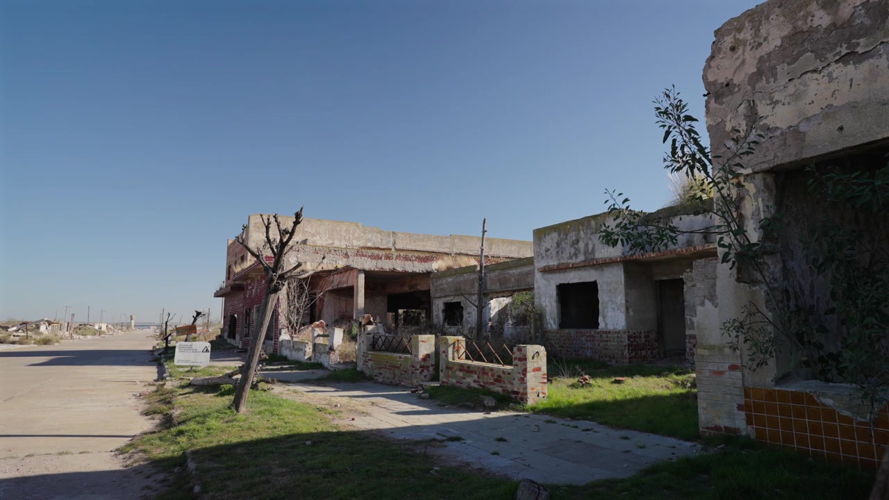 Una vista a livello stradale delle desolate e abbandonate rovine di cemento e degli alberi morti che costeggiano una strada asfaltata nella famosa città fantasma di Villa Epecuén, Argentina.